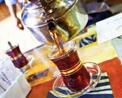 Here's a one-sentence description of the image:A metal teapot pours dark liquid, likely tea, into a clear, ornate glass cup sitting on a saucer, with another cup visible in the background on a patterned surface.