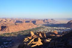 An elevated view reveals a desert landscape with rugged red rock formations, a lush green valley, and a densely packed town nestled within the arid terrain under a clear blue sky.