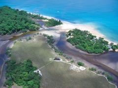 An aerial view shows a tropical coastline with lush green forests, sandy beaches, and calm blue waters, with a small boat visible in the distance.