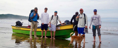 A group of six men and a dog stand in front of a bright yellow and green boat on a beach with calm water and overcast skies.