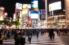 A bustling night scene in Shibuya Crossing, Tokyo, with crowds of people crossing the street amidst towering buildings adorned with bright advertisements and signage.