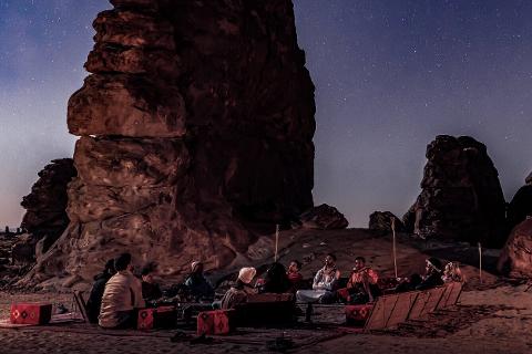 A group of people sits on a desert floor with rugs and cushions, surrounded by large rock formations under a starry night sky.