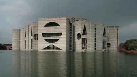 The Jatiya Sangsad Bhaban, the National Assembly Building of Bangladesh, stands in the middle of a pool of water under a cloudy sky.