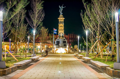 A nighttime view of a monument topped with an angel statue, framed by trees and illuminated by streetlights, with a paved walkway leading towards it.