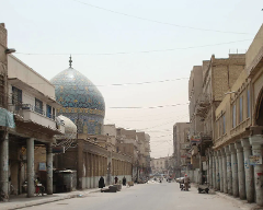 A street view in an old city, possibly in the Middle East, featuring weathered buildings with columns and a large, ornate blue-tiled dome in the background.
