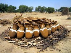 A large pile of brown, handmade pots with white heart-shaped designs are stacked on sticks and dry grass, set against a dry, rural landscape with scattered trees.
