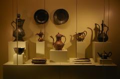 This is a display of antique metal objects, including pitchers, plates, a bowl, a box, knives, and a teapot, arranged on white pedestals in a museum setting.