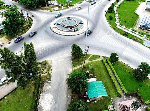 Aerial view of a roundabout with a fountain, surrounded by green lawns and busy roads.