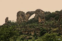 A scenic landscape features rock formations with a natural arch and lush greenery under an overcast sky.