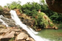 A scenic view of a tiered waterfall cascading into a green pool surrounded by rocks, trees, and vegetation.