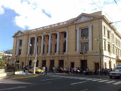 A bustling street scene in front of the Teatro Nacional, featuring pedestrians crossing the road and a taxi parked nearby, set against a backdrop of a grand building with columns and architectural details.