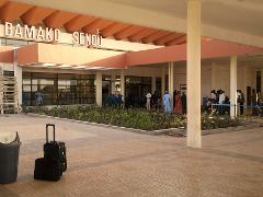 Mali Private Armored Car Hire and Armed Security Service A shot of the Bamako Senou Airport in Mali, showcasing a building with a sign, some people waiting outside, luggage, and greenery.
