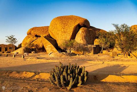A desert landscape featuring striking rock formations, a small building, and a clump of cacti in the foreground under a clear blue sky.