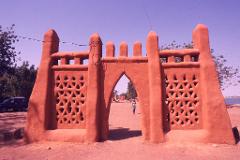 A person walks through a clay gate with triangular openings, set against a clear sky and sandy ground.