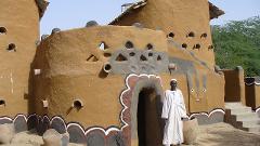 A man stands in front of a traditional mud-brick building with decorative openings and painted features.