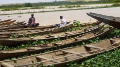 Niamey and its Exotic Surroundings 5-Day Several wooden boats are beached on the shore, some containing watermelons and two men. Vegetation and trees can be seen across the water.