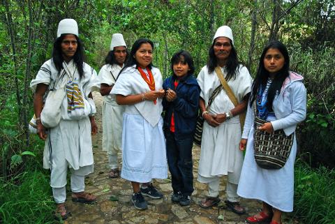 Katanzama Indigenous Community Hiking Tour with Poza Sagrada Visit A group of Indigenous people in traditional white clothing, standing on a stone path surrounded by lush green foliage.