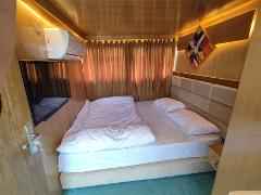 A cozy hotel room with a double bed, beige headboard, brown curtains, a TV, air conditioner, and colorful artwork on the wall. The room has wood paneling with soft, ambient lighting.