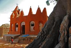 A child walks near an orange mud building with a large tree in the foreground, showcasing a traditional African village scene.