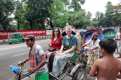 Two tourists are riding in a colorful rickshaw being pulled by a smiling man in Bangladesh, with other rickshaws and people nearby.