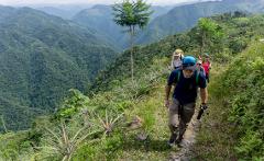 A group of hikers walks along a lush trail overlooking a deep valley filled with tropical rainforests, under a cloudy sky.