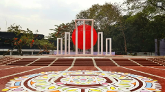 The Shaheed Minar, a national monument in Dhaka, Bangladesh, stands tall with its iconic white columns and a red sun backdrop, on a brick-paved plaza adorned with a vibrant, colorful rangoli design.