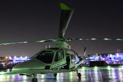 A helicopter labeled "Expo 2020 Dubai, UAE" sits on a landing pad at night, illuminated with green lights and reflecting on the wet surface.