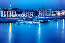 Here's a description of the image:A nighttime view of a harbor with boats docked along the waterfront, illuminated by city lights reflecting on the calm water.