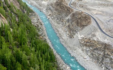 A turquoise river flows between a lush green forest and a rocky, barren landscape with a winding road.