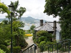 A charming garden pathway leads down stairs, past lush greenery and a quaint house with a tiled roof, offering a scenic view of a city and mountain backdrop.
