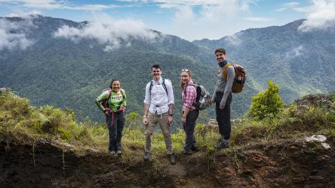 Four hikers stand on a rocky ledge with a mountainous, lush, and partially cloudy landscape behind them.