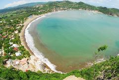 A panoramic aerial view shows a beautiful bay with turquoise water, a sandy beach lined with buildings and trees, and lush green hills in the background in Nicaragua.