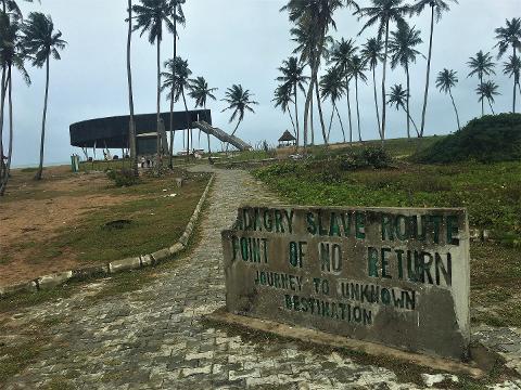 A stone sign reading "Badagry Slave Route - Point of No Return" stands at the start of a path leading towards a beach with palm trees and a modern structure.