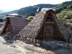Two small, A-frame huts with thatched roofs stand on a rocky ground, with a mountainous landscape in the background.
