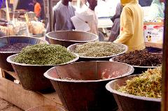A vibrant market scene featuring large bowls filled with a variety of herbs and spices, with people and other merchandise visible in the background.