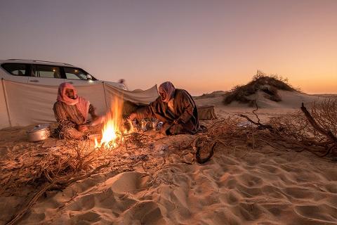 Two men in traditional Arabian attire are camped in the desert, tending to a fire with tea pots, with a vehicle and tent visible behind them against the warm sunset.