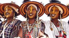 Three men with dark skin and yellow face paint wear traditional clothing and hats in a cultural festival.