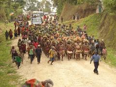 A vibrant procession of people, some in traditional attire, moves along a dirt road in Papua New Guinea, with vehicles and green hills in the background.