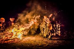 A vibrant nighttime scene captures a fire dance performance with performers in traditional masks and costumes, with a large bonfire and sparks creating an intense atmosphere.