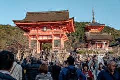 A crowd of people gathers in front of the Kiyomizu-dera temple in Kyoto, Japan, with the iconic red pagoda and gate visible in the background.