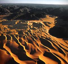 This aerial shot showcases the striking contrast between golden sand dunes and dark, rugged mountains in the Aïr Mountains, highlighting the harsh yet beautiful landscape.