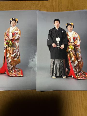 A Japanese couple in traditional wedding attire poses against a gray backdrop; the bride in a vibrant kimono adorned with cranes and flowers, and the groom in a black robe with hakama.
