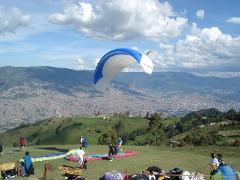A paraglider takes off on a sunny day from a grassy hill overlooking a sprawling city, while people prepare their equipment and relax on the ground.