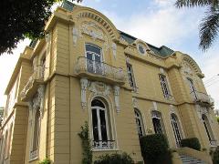 A yellow, ornate, three-story building stands tall, featuring arched windows, balconies, and intricate white detailing against a partly cloudy sky.