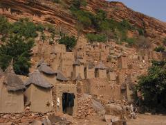 A Dogon village in Mali nestled at the foot of a cliff, featuring mud-brick houses with thatched roofs and rocky terrain.