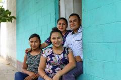 A family of four poses in front of a turquoise-colored brick building, with two daughters seated in front and parents standing behind them in a doorway.