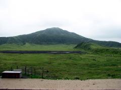 A wide, green landscape with a dark lake in the middle, surrounded by hills and mountains under a cloudy sky.