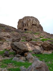 A landscape featuring a large, craggy rock formation with a cave-like opening, surrounded by scattered rocks and patches of green grass under an overcast sky.
