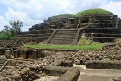 A stepped pyramid with stone stairs and a small building at the top, possibly an ancient Mesoamerican structure, under a cloudy sky.