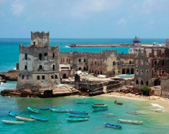This is a coastal view of a historic, weathered building complex, featuring a prominent tower and numerous boats dotting the turquoise waters.